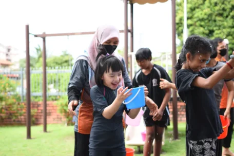 a smiling child holding up a small blue bucket in a playground